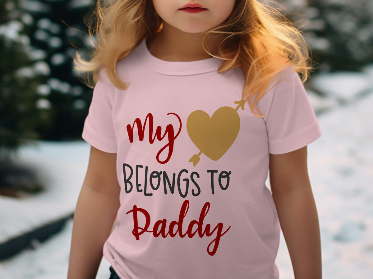 A young child wearing a light pink t-shirt with the text My Belongs to Daddy and a gold heart graphic.