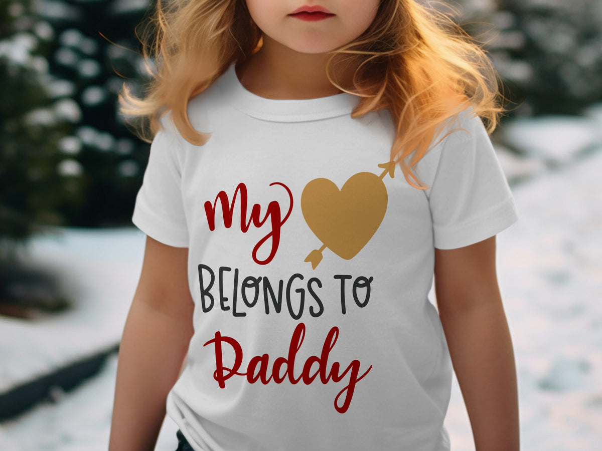 A child wears a white t-shirt printed with red and black text reading My Belongs To Daddy and a gold heart pierced by an arrow.