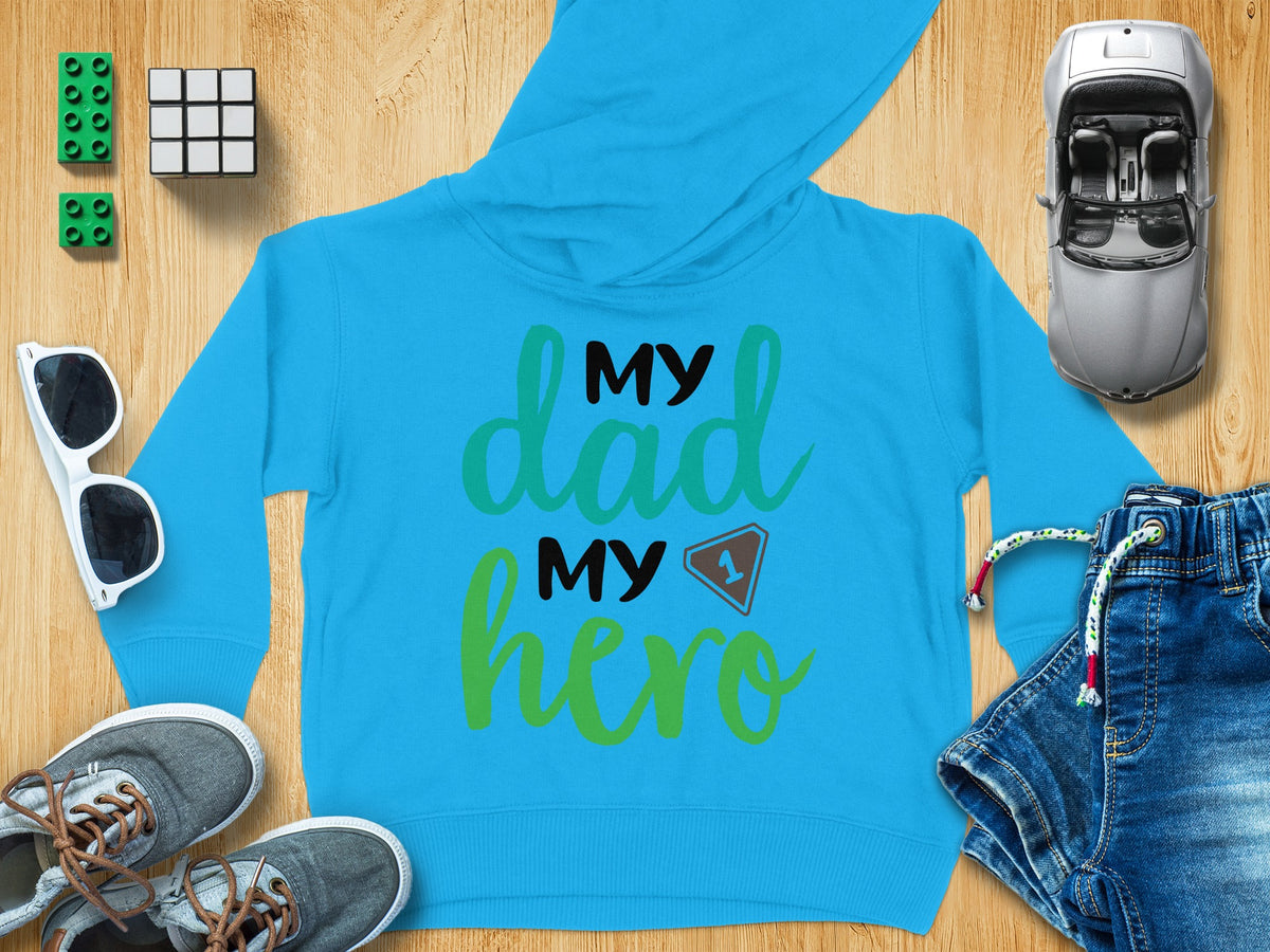 A bright blue children's hoodie featuring the text My Dad My Hero, surrounded by toys and clothing items on a wooden background.