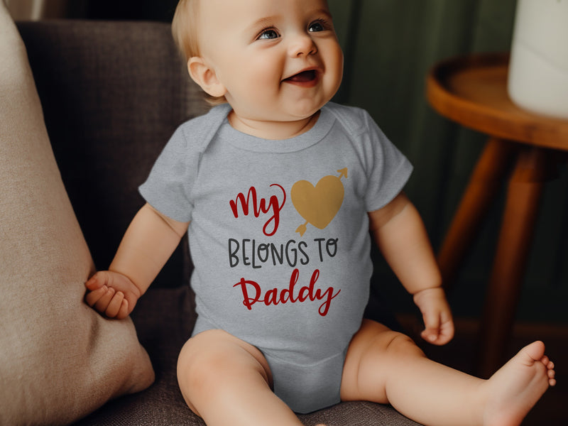 A smiling baby wears a grey bodysuit printed with the red and gold text "My [heart icon] Belongs to Daddy."