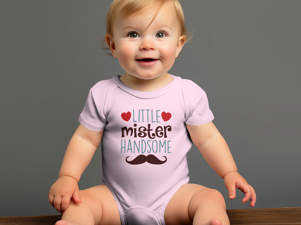 A smiling baby wearing a light pink onesie printed with the text Little Mister Handsome and a mustache.