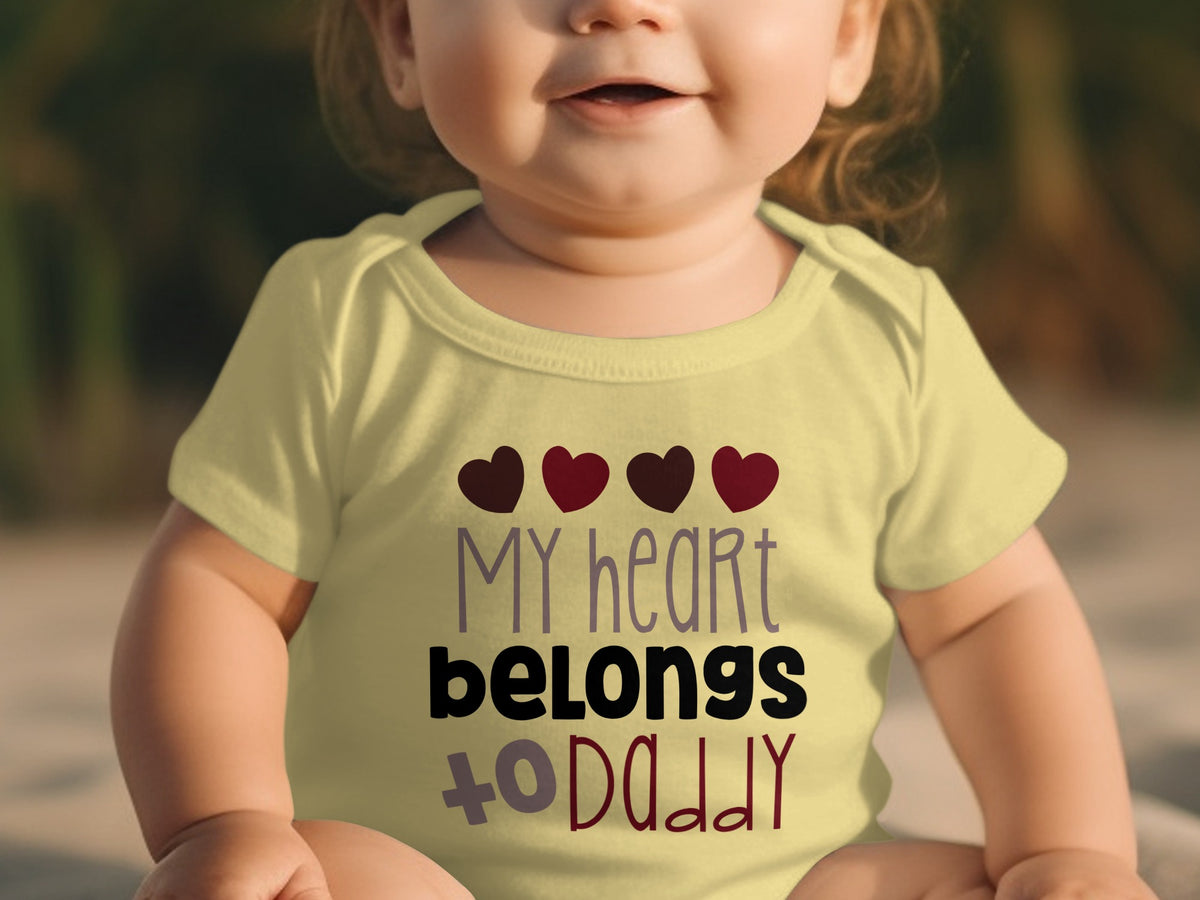 A smiling baby wearing a light yellow onesie with the text My heart belongs to daddy.