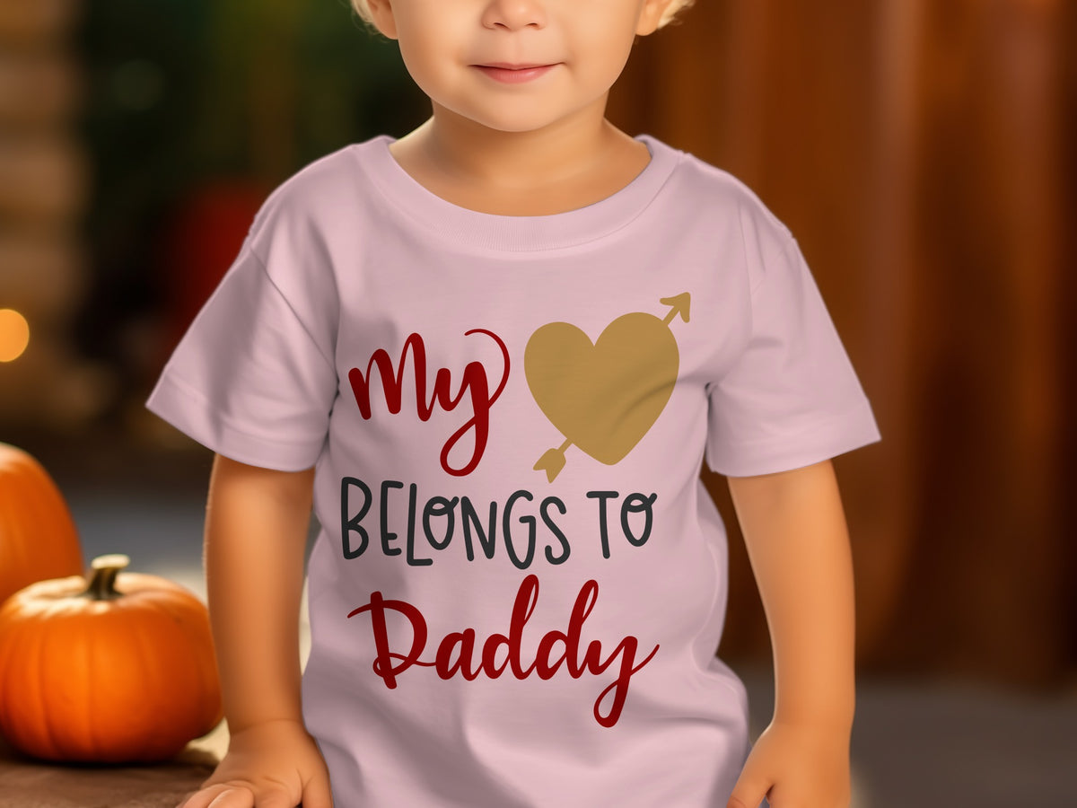 A toddler wears a pink t-shirt printed with the text My Belongs to Daddy and a gold heart pierced by an arrow.