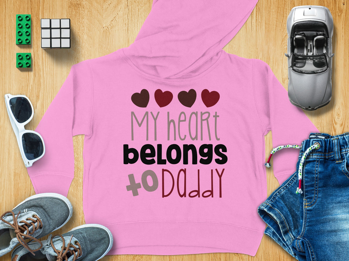 Pink children's hoodie with the text "MY heart belongs to DADDY" and four hearts, surrounded by toys and accessories.