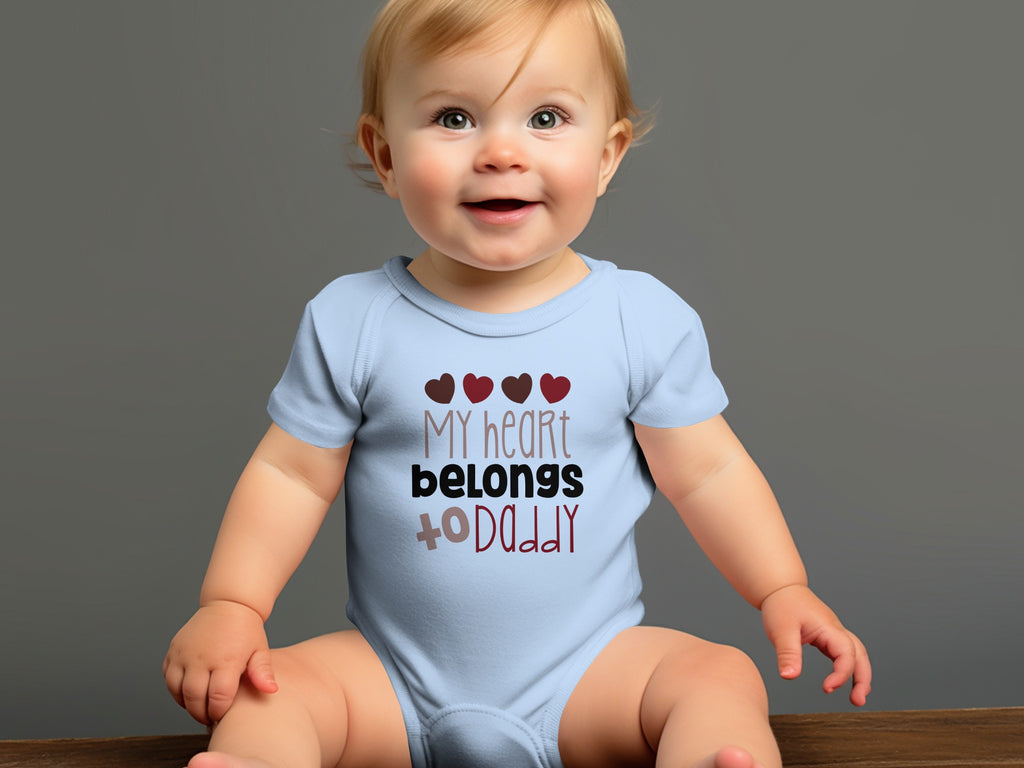 A smiling baby sits wearing a light blue bodysuit with the text My heart belongs to Daddy.