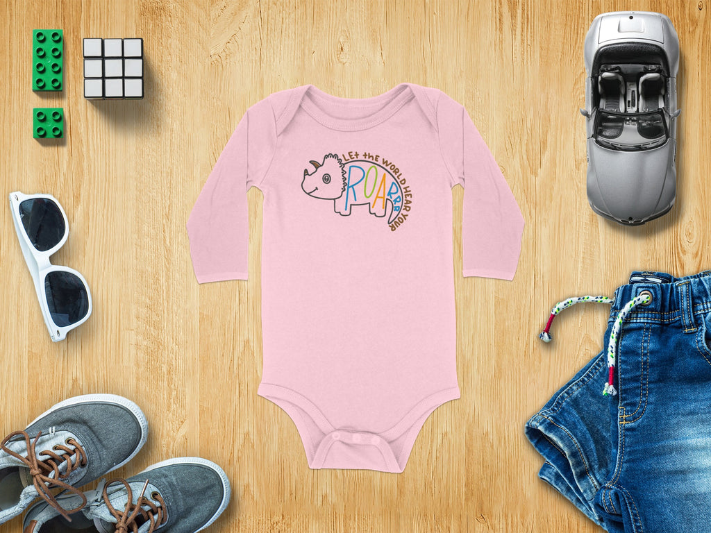 A pink long-sleeved baby bodysuit with a triceratops graphic is centered on a wooden background, surrounded by toys and clothing.