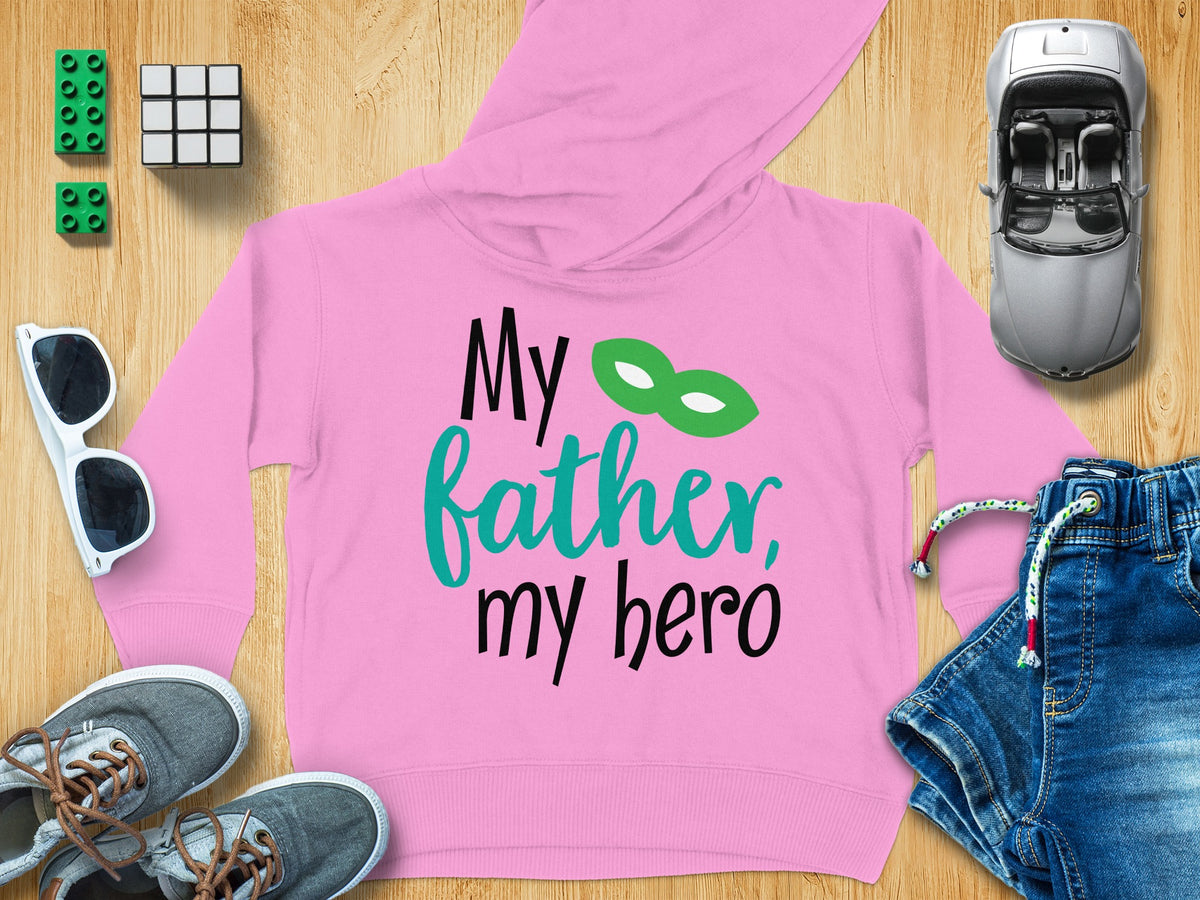 A pink children's hoodie with black and teal text reading "My father, my hero" surrounded by toys on a wooden surface.