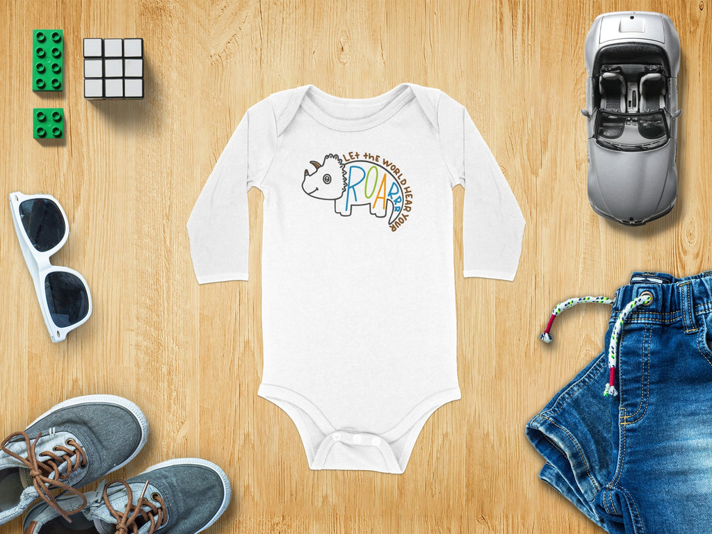 A white long-sleeved baby bodysuit with a triceratops graphic lies flat on a wooden surface surrounded by casual accessories and denim shorts.