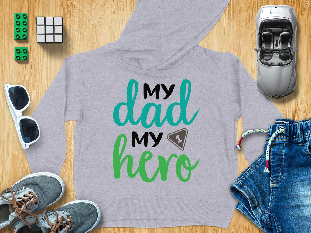 Grey children's hoodie with the black, teal, and green text My Dad My Hero, surrounded by toys and clothes on a wooden background.