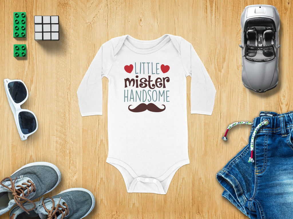 White long-sleeve baby onesie printed with Little Mister Handsome and a mustache, surrounded by toys and clothing on a wooden background.