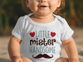 A toddler wearing a white short-sleeve bodysuit featuring the text little mister handsome and a mustache graphic.