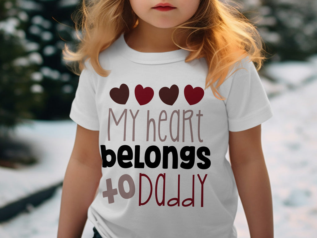 A young blonde child wears a white t-shirt printed with the text MY heart belongs to DADDY and four hearts.