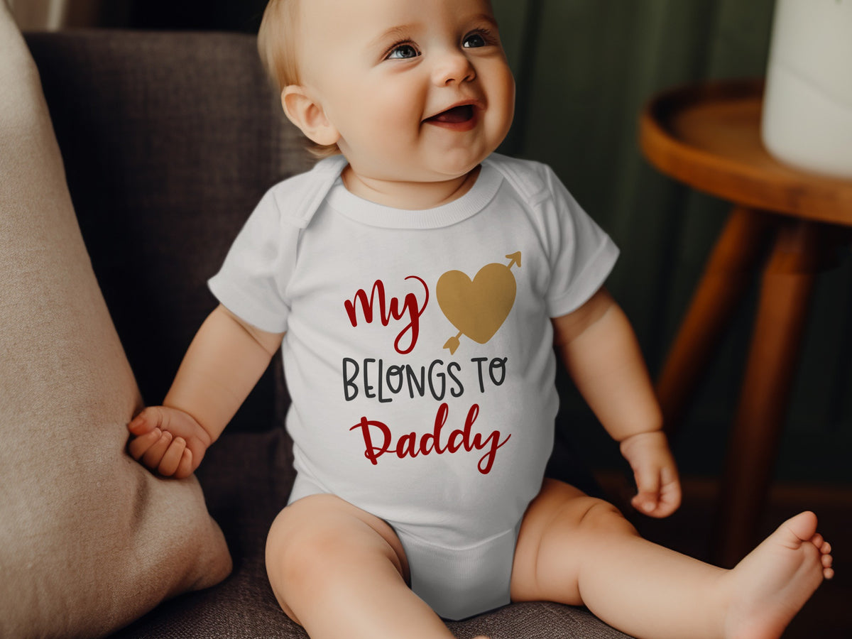 A smiling baby wearing a white bodysuit with red and black text reading My Heart Belongs to Daddy.