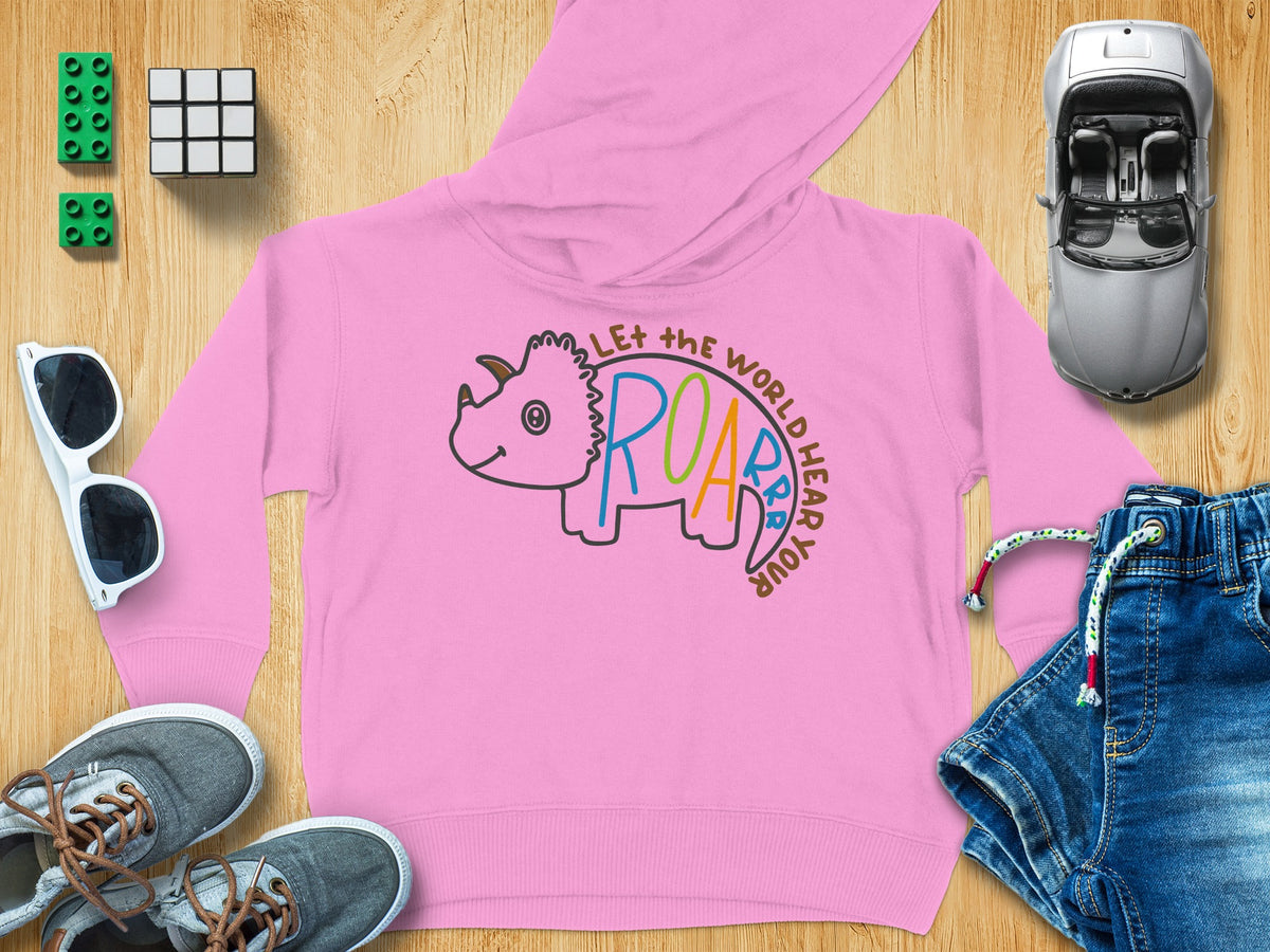 A pink children’s hoodie with a triceratops graphic reading Let the world hear your roar, surrounded by toys and clothing on wood.