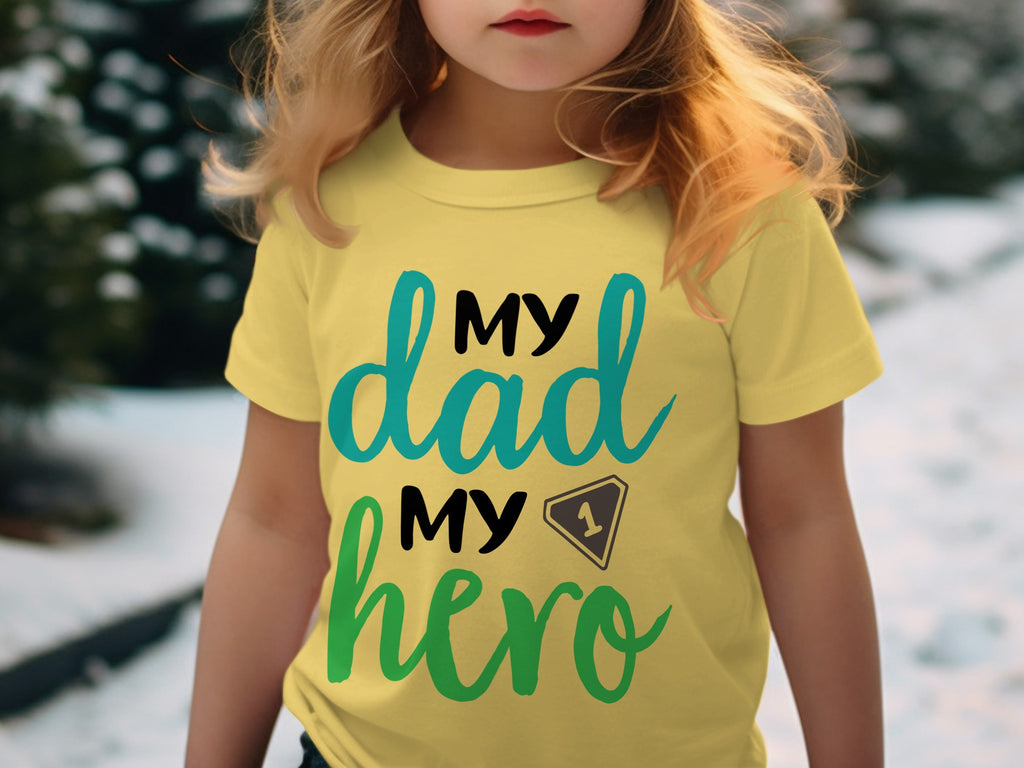 A young child wearing a yellow t-shirt with black, turquoise, and green lettering that reads My dad, my hero.