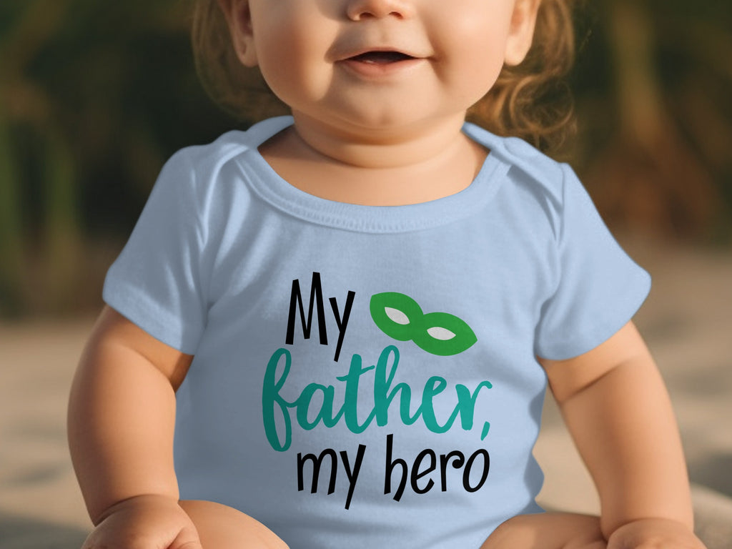 A baby wears a light blue onesie printed with the text My father, my hero and a green superhero mask.