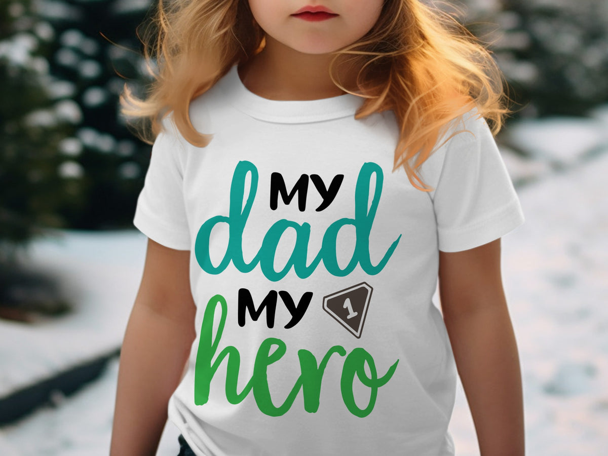 A young child with blonde hair wears a white t-shirt printed with the text My dad My hero and a number one graphic.