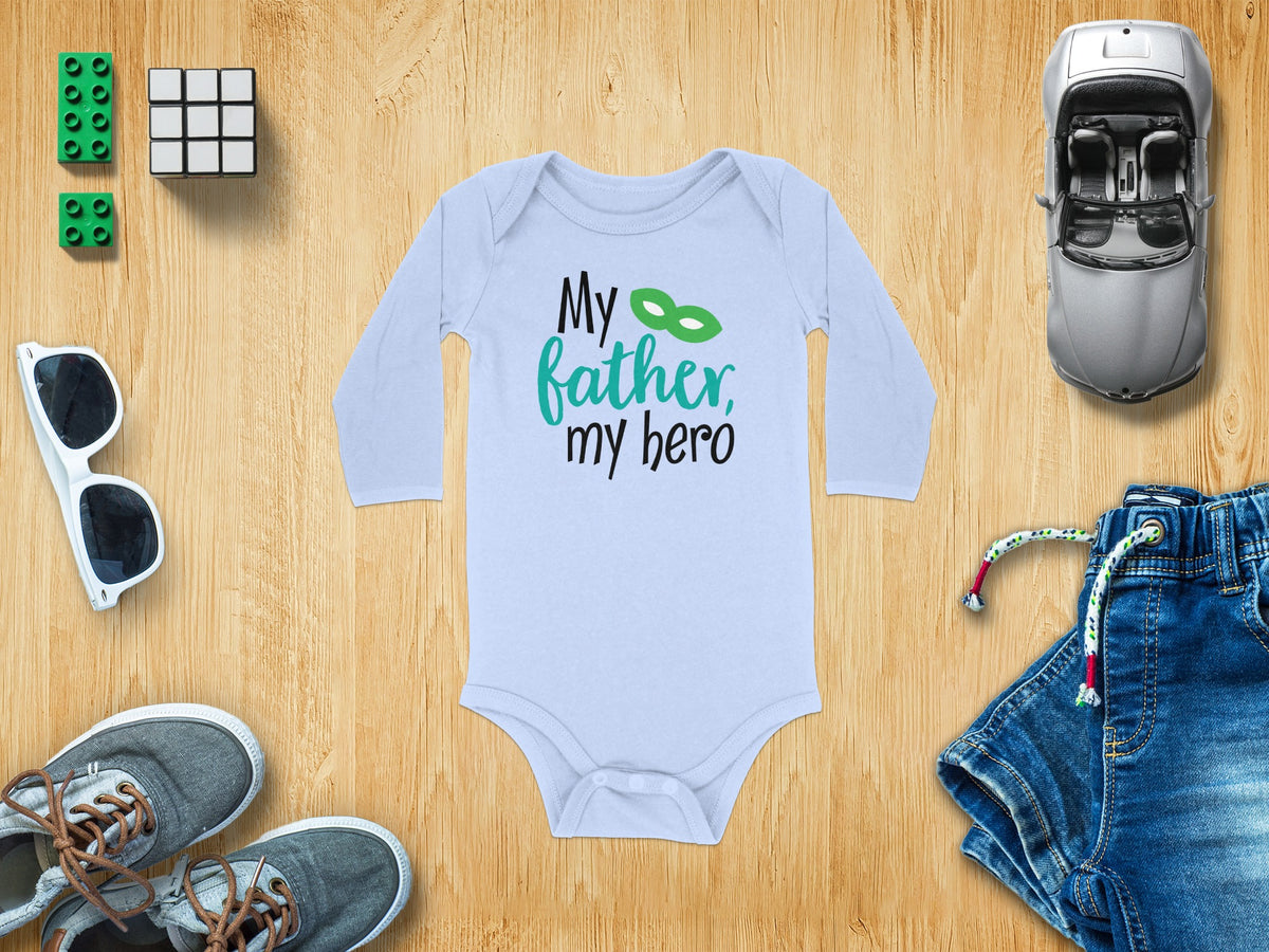 Light blue long-sleeve baby bodysuit with black and green text reading My father, my hero, surrounded by toys on a wooden background.