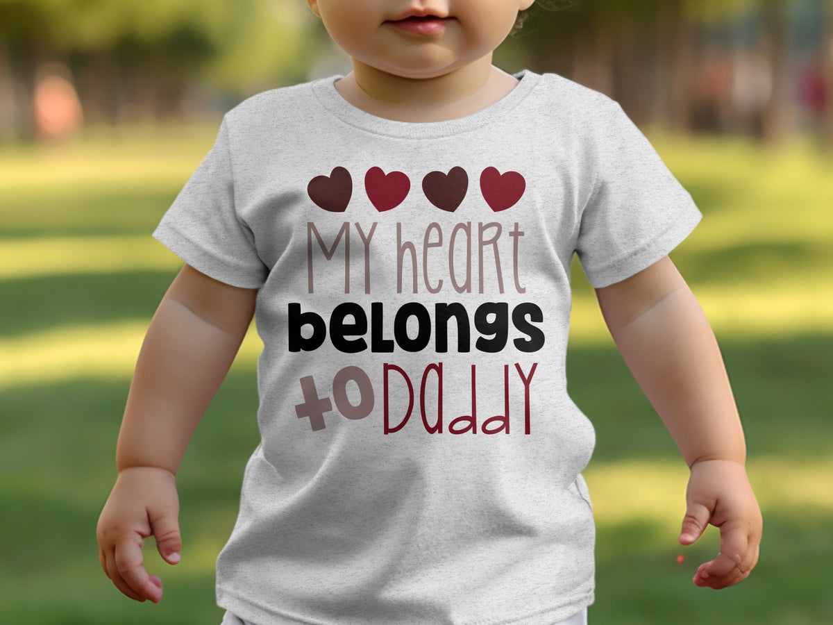A toddler wearing a light grey t-shirt featuring the text "my heart belongs to daddy" and four small hearts.
