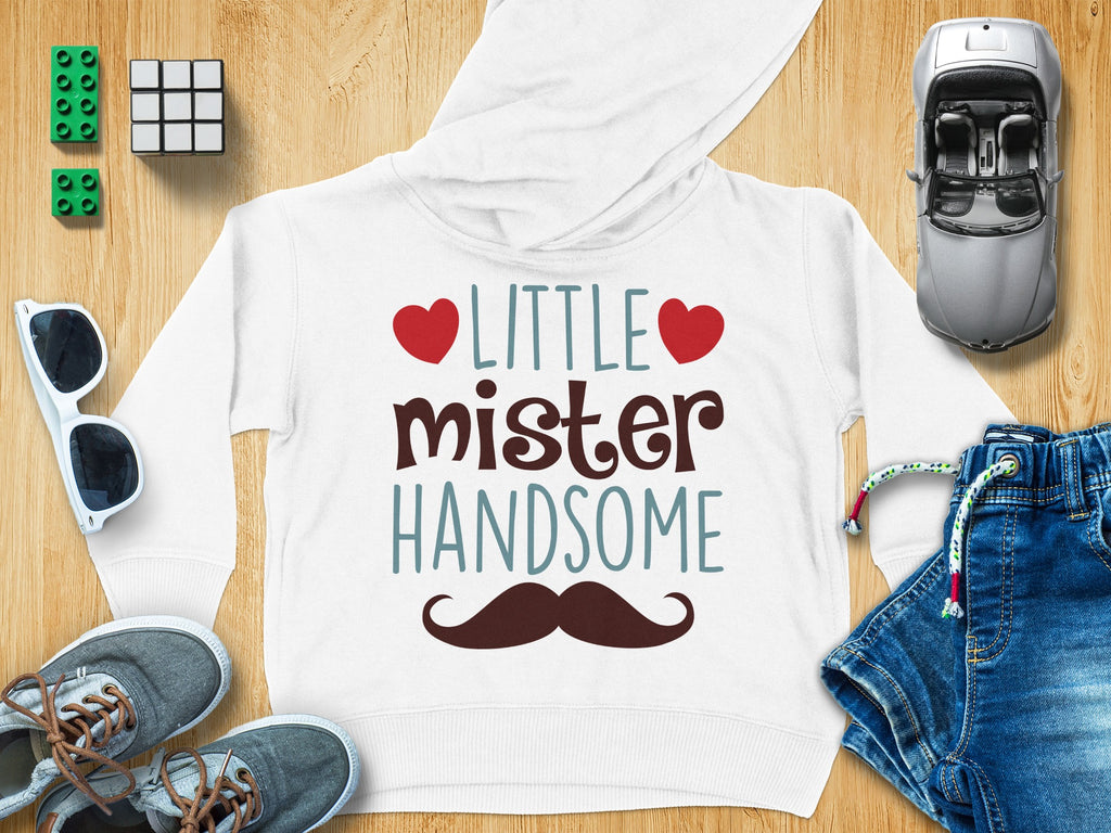 A white children's hoodie with the text "LITTLE mister HANDSOME," red hearts, and a brown mustache, surrounded by toys and clothing on a wooden surface.