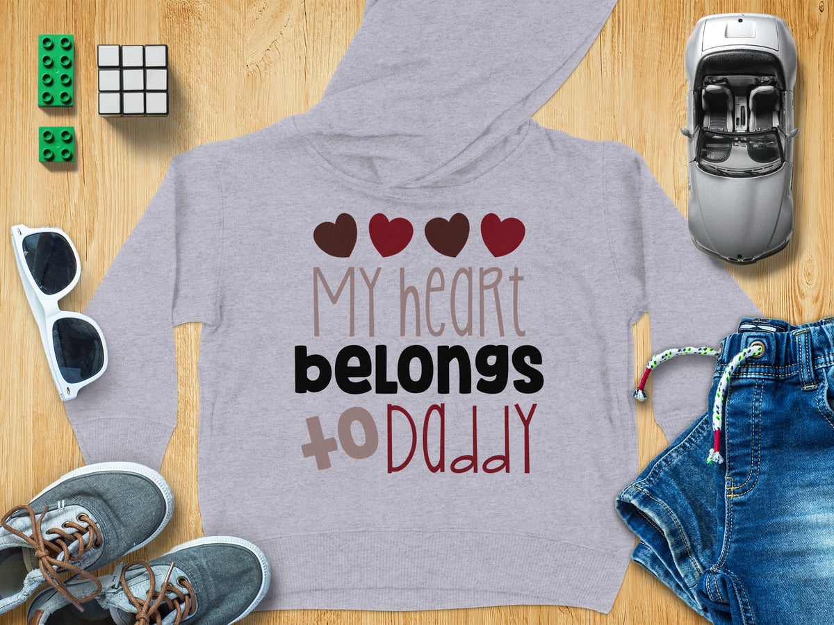 A grey children's hoodie with the text My heart belongs to Daddy surrounded by toys on a wooden surface.