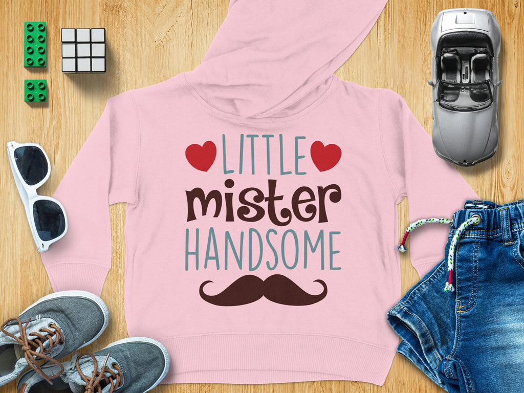 A light pink children's hoodie with the text LITTLE mister HANDSOME and a mustache graphic, surrounded by toys and clothes on a wooden background.