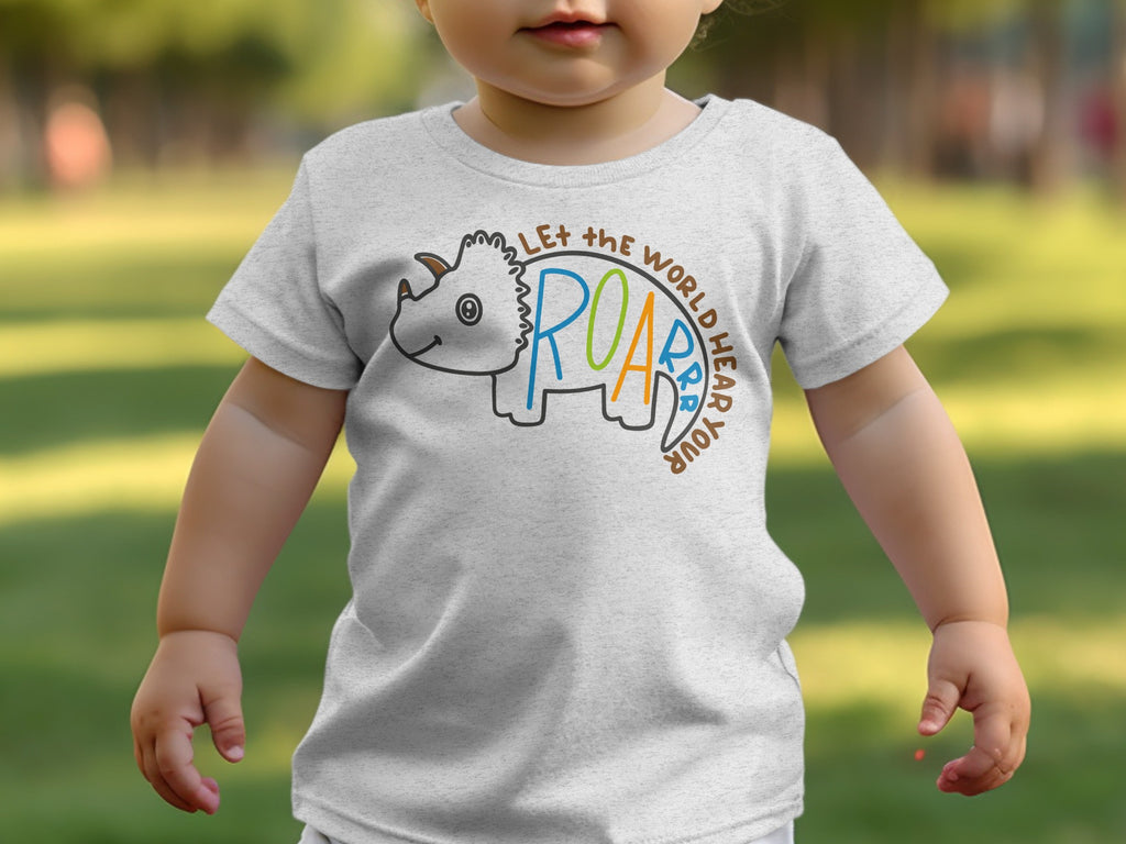 A toddler wearing a light grey t-shirt featuring a triceratops graphic and the text "Let the world hear your roar."