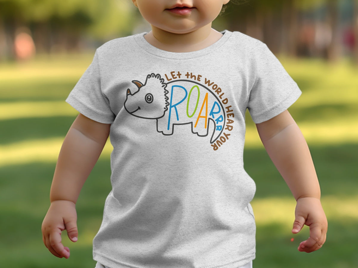 A toddler wearing a light grey t-shirt featuring a triceratops graphic and the text "Let the world hear your roar."
