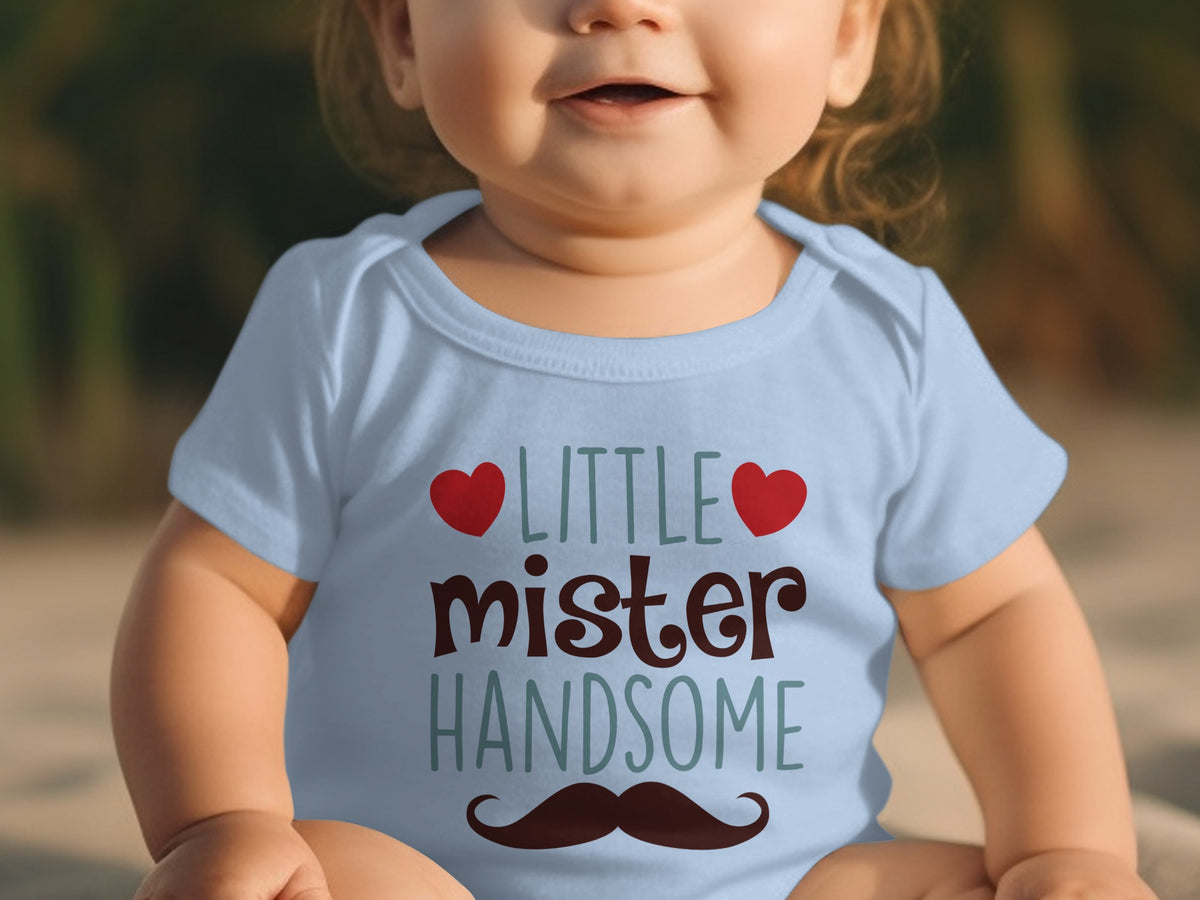 A baby wearing a light blue onesie printed with the text little mister handsome flanked by red hearts.