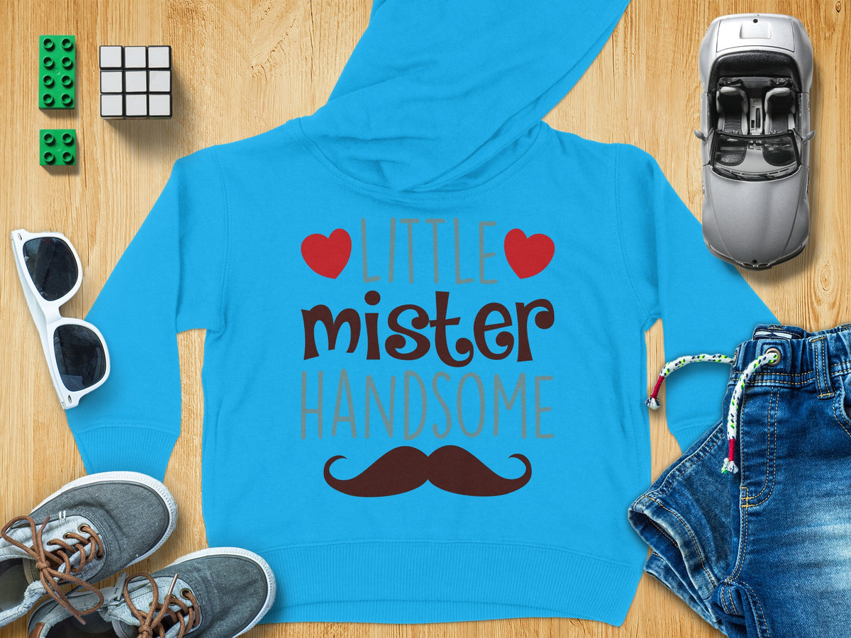 Bright blue children's hoodie with the text little mister handsome and a brown mustache, surrounded by toys, sunglasses, shoes, and jeans on a wooden surface.