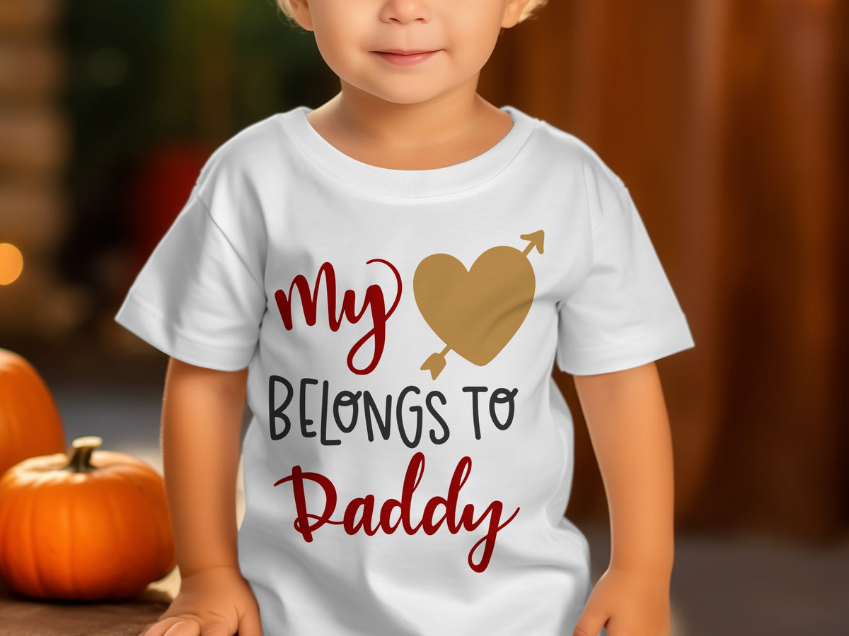A toddler wears a white t-shirt featuring the printed text My Belongs to Daddy and a gold heart pierced by an arrow.