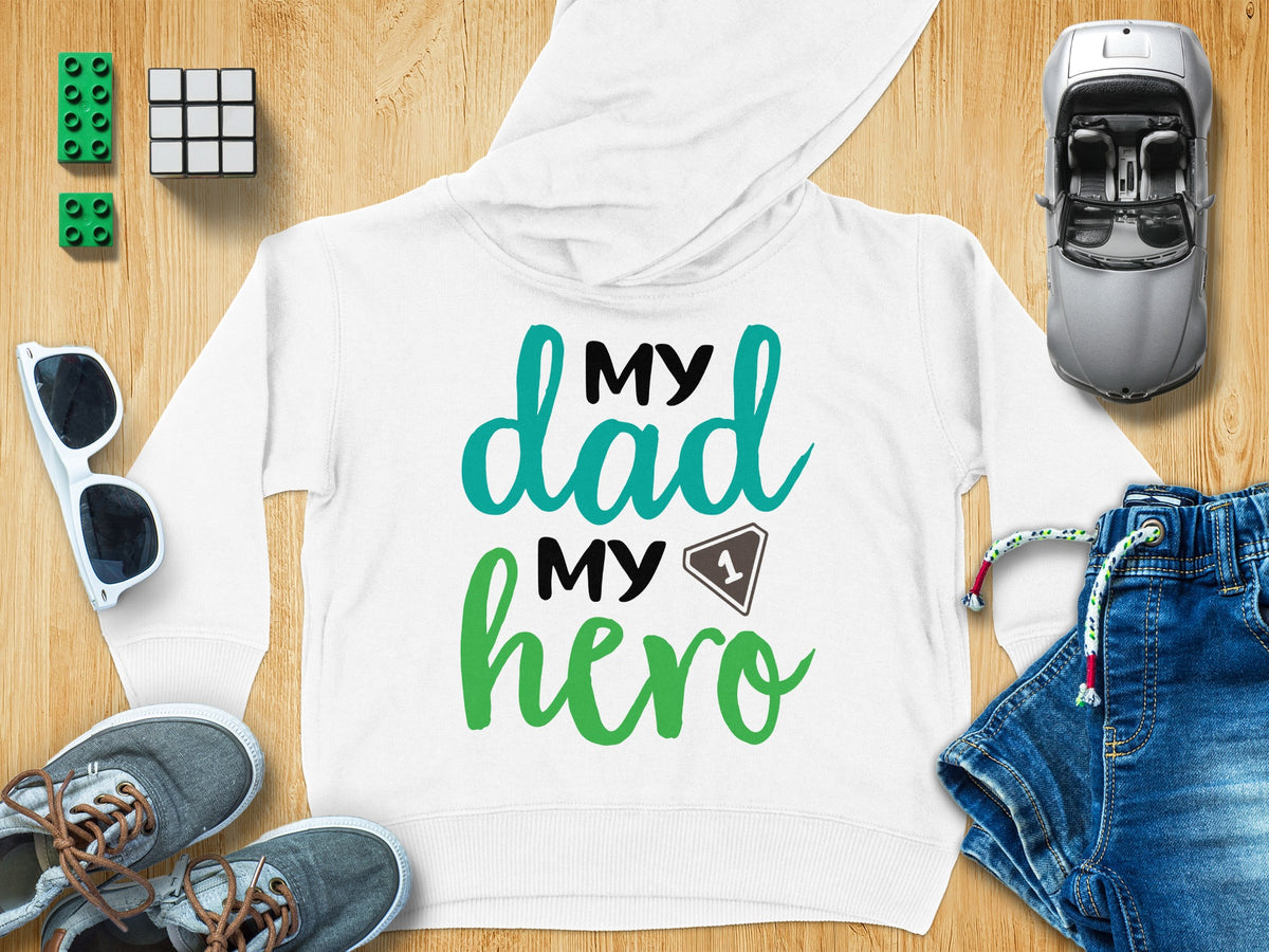 A white children's hoodie featuring the text My dad My hero surrounded by toys and accessories on a wooden surface.