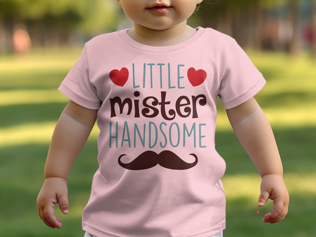 A toddler wearing a pink t-shirt printed with the text LITTLE mister HANDSOME and a mustache.