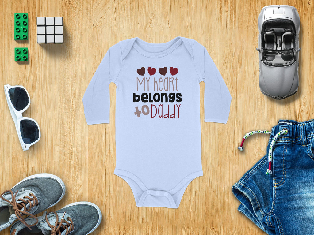 Light blue long-sleeve baby bodysuit with text "My heart belongs to Daddy" surrounded by toys on a wooden surface.
