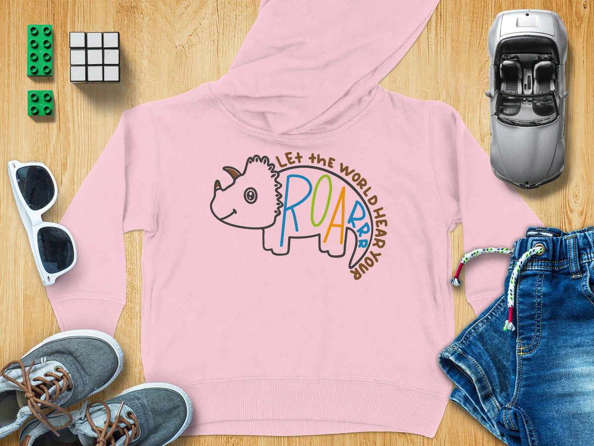 Pink children's hoodie with a triceratops graphic and text, surrounded by toys and clothing on wood.