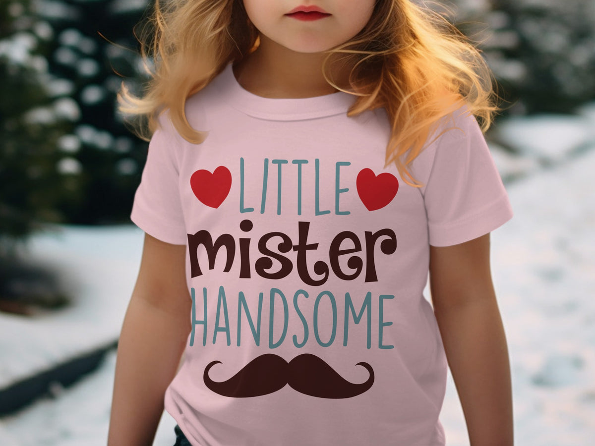 A young child wearing a light pink t-shirt with the text Little Mister Handsome, red hearts, and a mustache graphic.