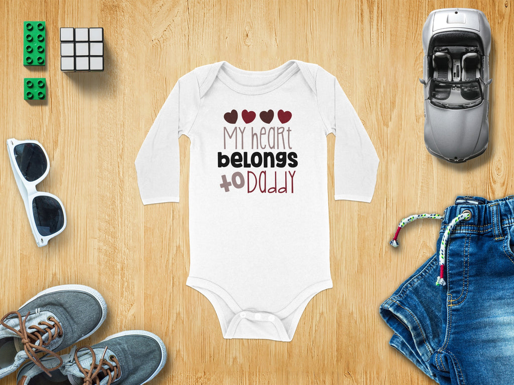 White long-sleeve baby bodysuit with My Heart Belongs to Daddy text, surrounded by toys and clothes on wood.