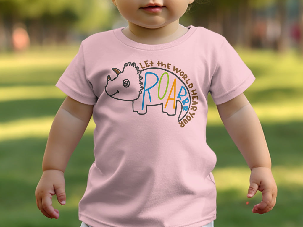 A toddler wearing a light pink t-shirt with a triceratops outline and text reading Let the world hear your roar.
