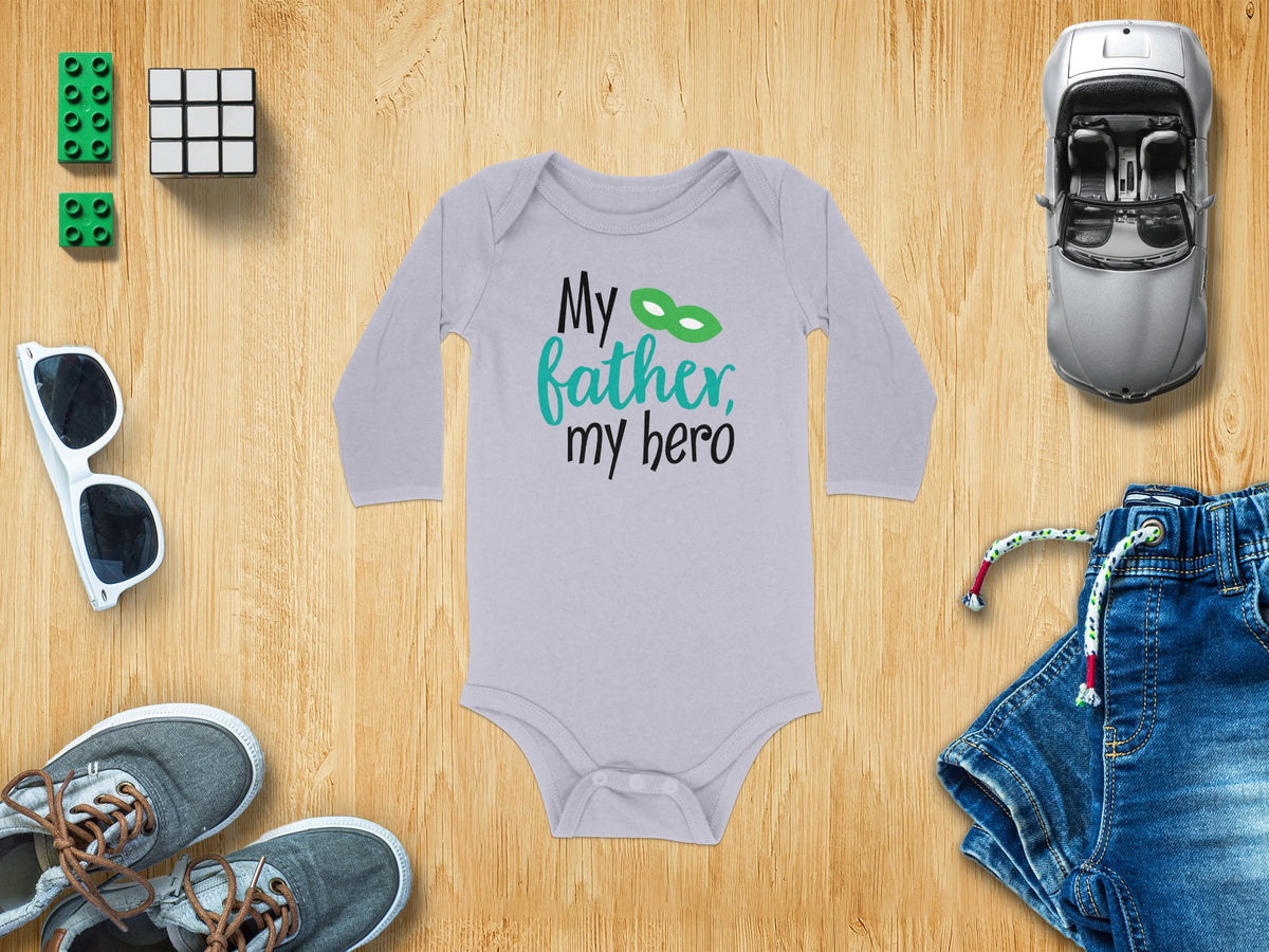 Light gray long-sleeve baby bodysuit with black and green text reading My father, my hero on a wooden background surrounded by toys and clothes.