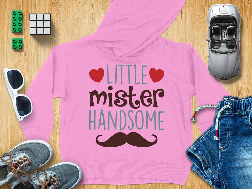 Pink children's hoodie with the text LITTLE mister HANDSOME, red hearts, and a brown mustache, surrounded by toys and accessories on a wooden background.