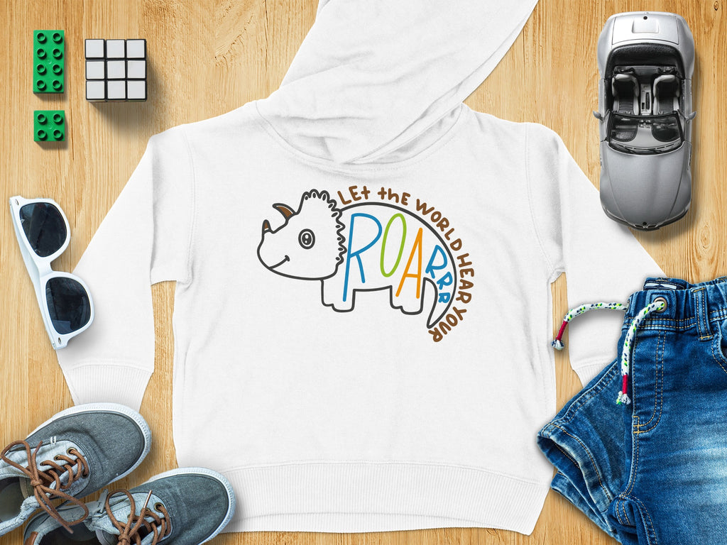 White children’s hoodie featuring a triceratops outline and text reading Let the World Hear Your Roar, surrounded by toys on a wooden surface.