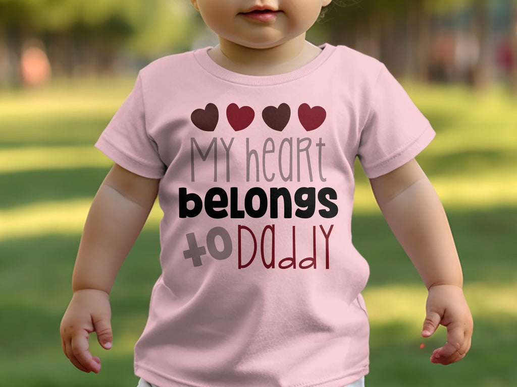 A toddler wears a pink t-shirt printed with the text "my heart belongs to daddy" and four hearts.