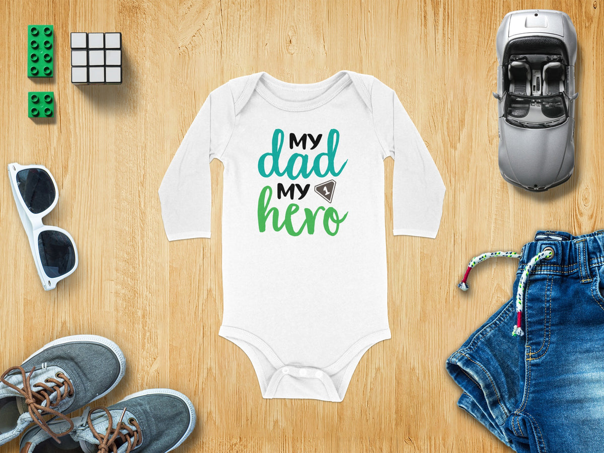 White long-sleeve baby bodysuit with My Dad My Hero text surrounded by toys on wood.