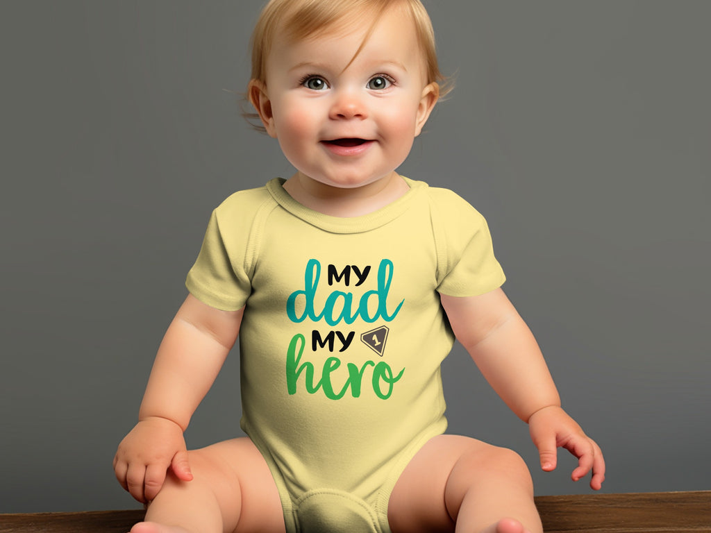 A smiling baby wearing a yellow short-sleeved bodysuit with the text My Dad My Hero printed on the chest.