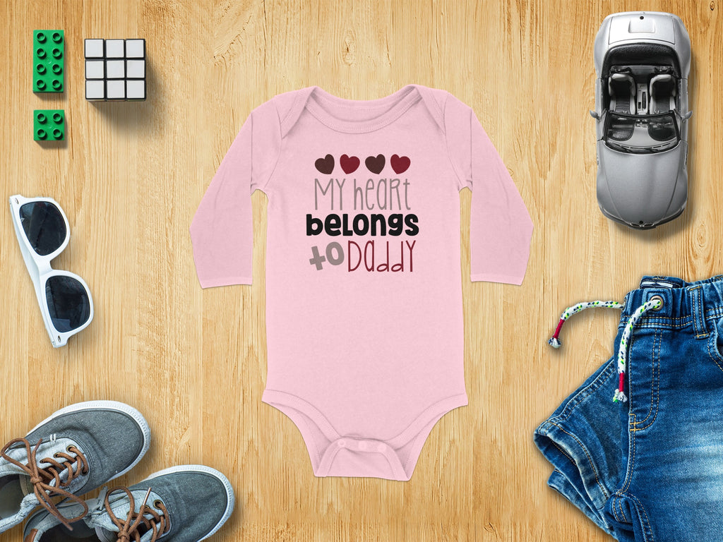 Pink long-sleeve baby onesie printed with My Heart Belongs to Daddy, surrounded by toys and clothing on wood.