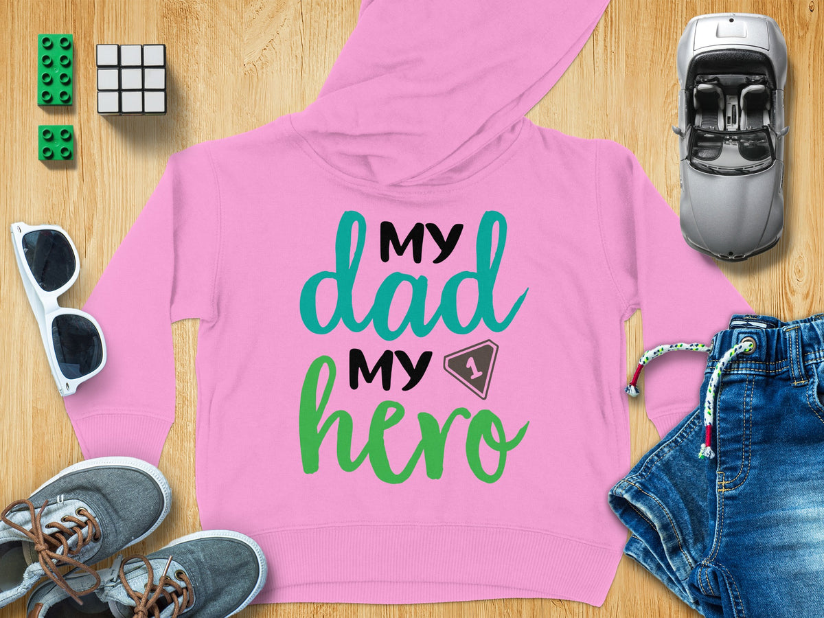 A pink hooded sweatshirt featuring the text My Dad My Hero, surrounded by toys and clothing items on a wooden background.
