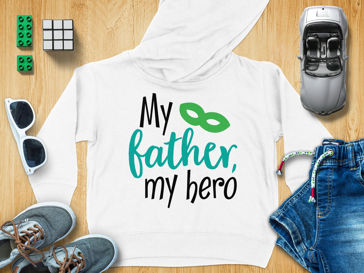 A white children's hoodie with black and teal text reading My father, my hero, surrounded by toys and accessories on a wooden background.