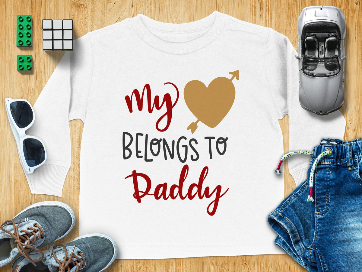 White long-sleeved children's t-shirt printed with the text My Belongs To Daddy surrounding a gold heart pierced by an arrow, displayed on a wooden surface with toys and denim jeans.