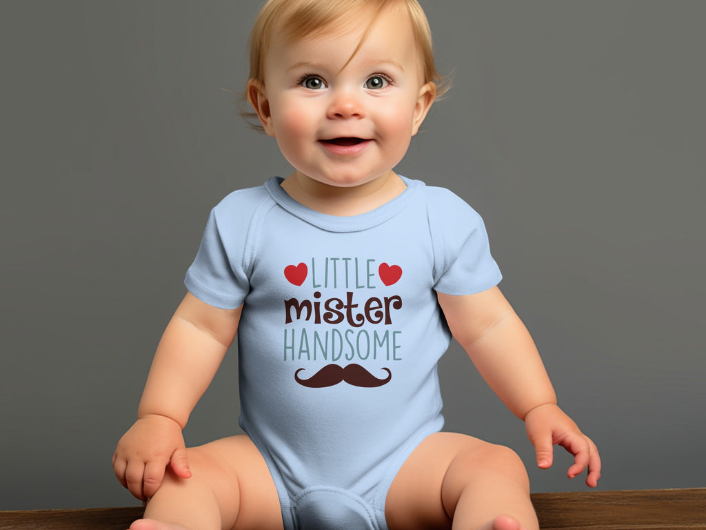 A smiling baby with blonde hair sits wearing a light blue onesie with text and a mustache graphic.