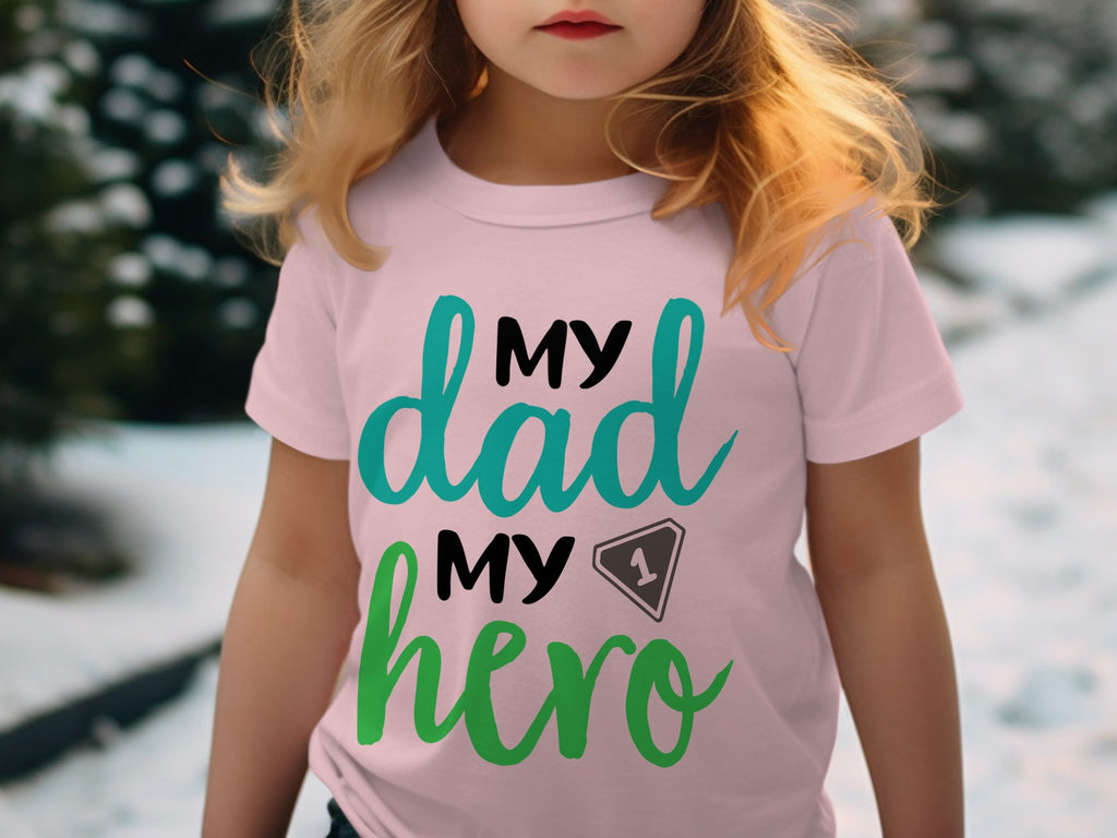 A child wearing a light pink t-shirt with black, turquoise, and green text reading My Dad My Hero.